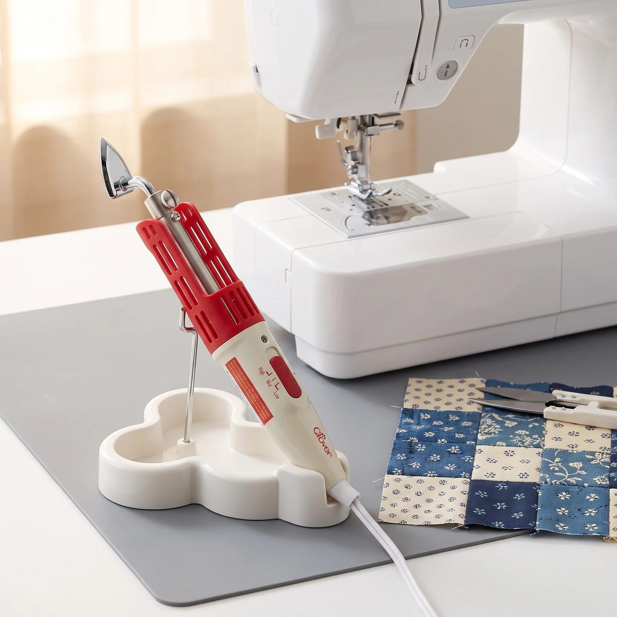Mini plancha Clover Mini Iron II en su soporte sobre un tapete de silicona, junto a una máquina de coser y un bloque de patchwork