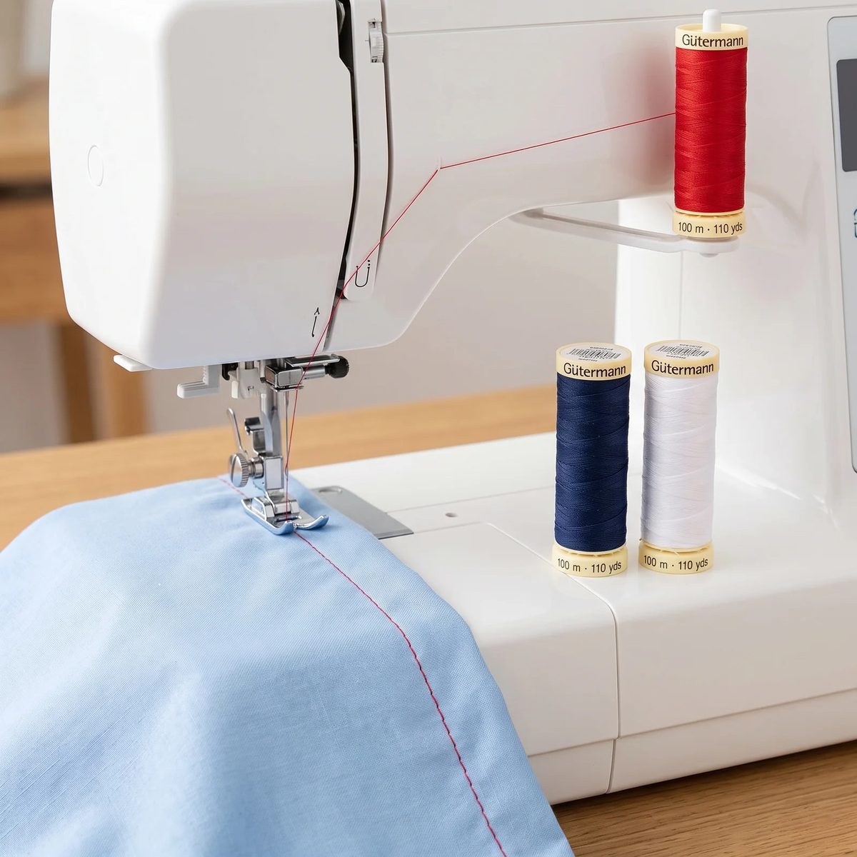 Carrete de hilo Gütermann Sew-All rojo montado en una máquina de coser casera, cosiendo una costura recta en algodón
