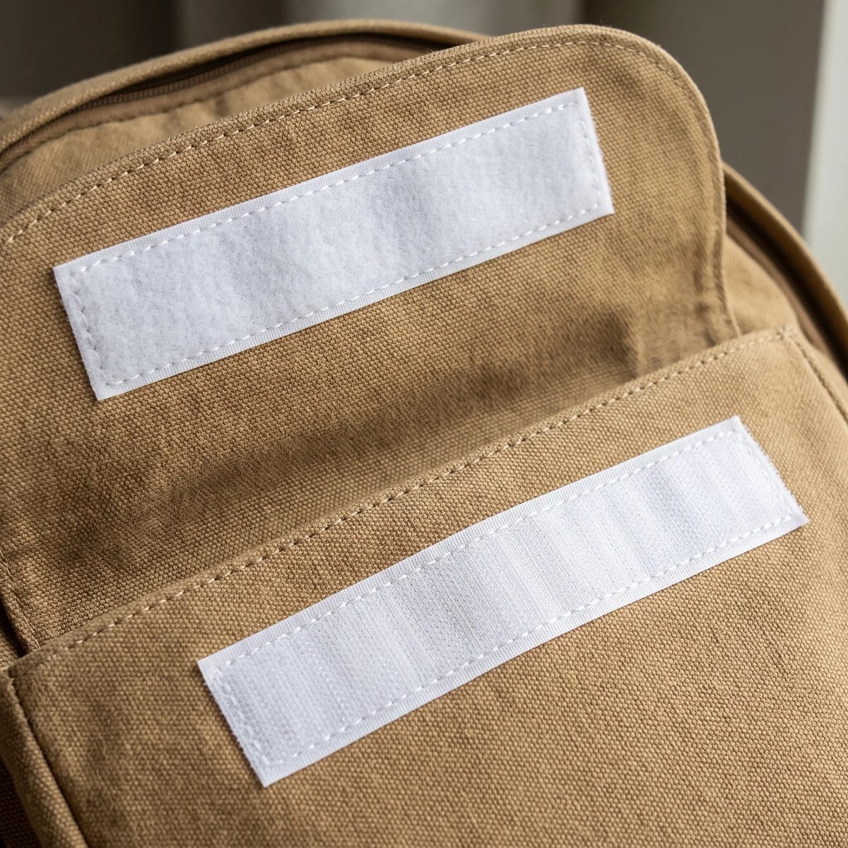 Dos tiras de velcro blanco cosidas en la solapa de un morral de lona, con la costura en cuadro visible